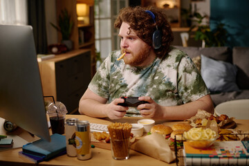 Man with curly hair wearing headphones and holding game controller while eating fries in cozy living room. Surrounded by fast food and desktop computer, focused on screen