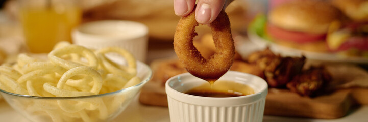 Hand holding onion ring dipping into sauce with curly fries, burger, and other snacks in...