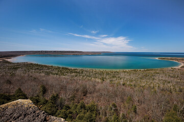In spring, an outlook above a bay of blue and turquoise waters along a rocky shoreline with forests and a rocky escarpment. A calm glass surface water under a bright blue sky with streaks of clouds. 