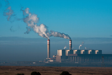 The Kendal Power Station stands prominently in an open landscape, with tall chimneys emitting smoke under a clear blue sky; sharp focus highlights the industrial structure against the natural surround