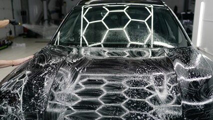 Professional car detailer applying soap and spreading foam across black car hood, meticulously...
