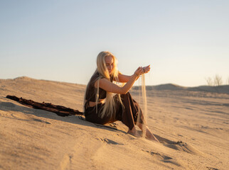 Woman Gently Pouring Sand Through Her Fingers While Sitting on Warm Desert Dunes During Sunset,...