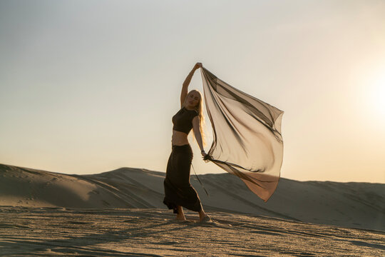 A woman stands in the desert, holding a light fabric poreo as the sun sets, casting long shadows over the sand dunes around her.