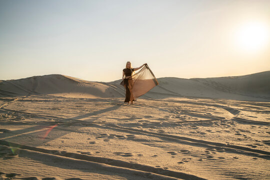 A woman stands in the desert, holding a light fabric poreo as the sun sets, casting long shadows over the sand dunes around her.