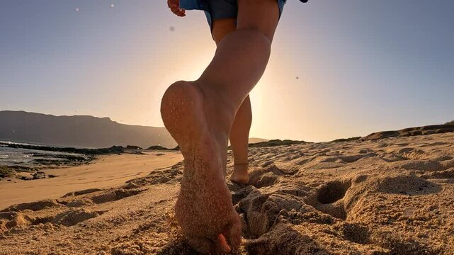 LOW ANGLE VIEW, SLOW MOTION, LENS FLARE: Barefoot woman runs across a sunlit beach, leaving clouds of sand behind. Carefree summer moment with young lady in skirt with shorts running in golden light.