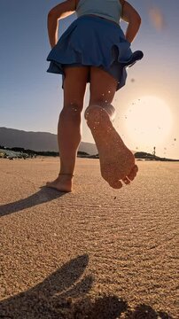 VERTICAL, LOW ANGLE VIEW, LENS FLARE: Fit young woman on a morning run along a sandy shore. At sunrise, she runs barefoot on beach in blue sportswear, leaving clouds of sand behind in golden light.