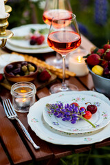 Beautiful elegant romantic dinner table decor, surprise day arrangement. Marriage proposal, engagement private party for two in a meadow. Sunset, golden hour. Cheese board, rose wine, fruits, lupine