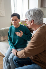 Tired nervous middle aged woman grabs chest struggles for air comforted on couch by supportive husband. Asthmatic episode, chest tightness, heart rhythm disruption, panic and fear of heart attack