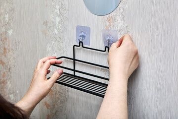 Person is pressing transparent adhesive hook onto bathroom wall while black metal shelf is already hanging on adjacent fixed hook.