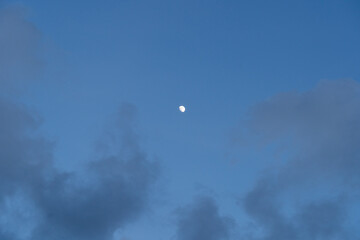 The moon shines brightly in tranquil blue evening sky, surrounded by faint, dark clouds creating peaceful atmosphere