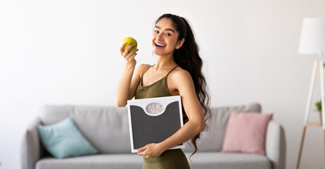 Plakat Portrait of millennial Indian lady holding scales and eating apple, choosing healthy diet, panorama. Young Asian woman preferring wholesome nutrition. Wellbeing and weight loss concept