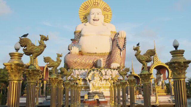 Giant laughing Buddha statue at Wat Plai Laem in Koh Samui, Thailand, surrounded by ornate golden pillars and reflecting in the temple tiled walkway. Travel and touristic landmark