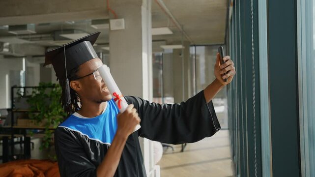 African American graduate in cap and gown celebrates achievement while talking to parents via smartphone video call