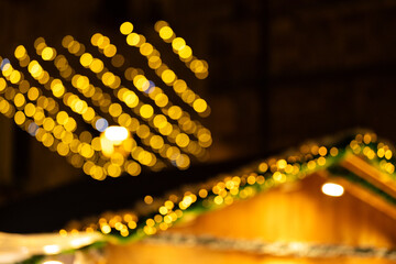 Defocused image of golden Christmas lights hanging above holiday market stall, creating warm and festive glow. The blurred bokeh effect enhances the holiday atmosphere