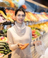 In vegetable store, young woman customer buy ripe carrot. Vegetables and fruits from different parts of world, products from local farms.