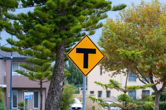 A yellow T-intersection street sign is located in a suburban residential neighborhood with trees and houses in the background, in Melbourne, Victoria, Australia.