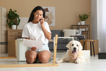 Young African-American woman with her cute dog assembling table and talking by mobile phone at home