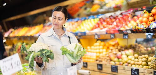 Positive middle-aged woman choosing cauliflower standing at counter in large vegetable market