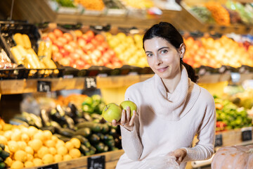 In vegetable store, young woman customer buy ripe apple. Vegetables and fruits from different parts of world, products from local farms.