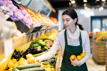 Vegetable shop assistant working in greengrocery. Woman seller in sales area with lemon in hands. Employee demonstrates supply of fruit. Local and imported farm vegetables and fruits