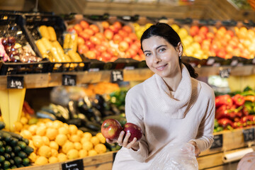 Near shelves and display cases in sales area of store, woman customer takes ripe apple out of box. Wide range of agricultural products. Self-service, comfortable shopping.
