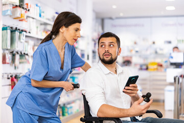 Obraz premium Positive woman pharmacist helping handicapped man in wheelchair choosing medical supplies at drugstore