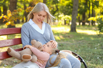 Cute little girl with toy bear and her mother resting on bench in park