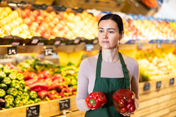 Polite middle-aged female seller offering red pepper standing by counter in large grocery store
