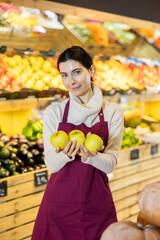 Woman staff with apple in hands, vendor demonstrate, adds and displays goods. In sales area of vegetable store, seller restructures display case.