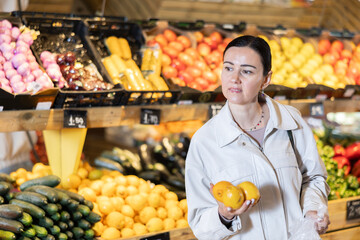 Positive middle-aged female buyer holding lemons in large fruit and vegetable market