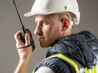 Construction worker wearing a white hard hat and safety vest communicates via handheld radio during site operations. Activity: Construction, Security, Site Management. © Grispb