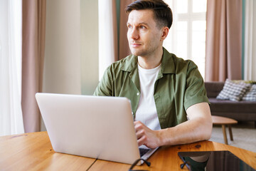 Thoughtful man works intently on laptop at wooden desk in bright, modern living room during a sunny afternoon