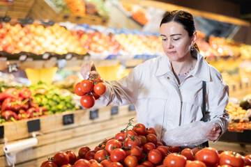Glad middle-aged female buyer purchasing tomatoes in grocery store with large assortment