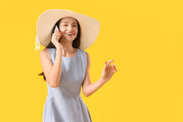 Beautiful young woman in hat talking by mobile phone on yellow background