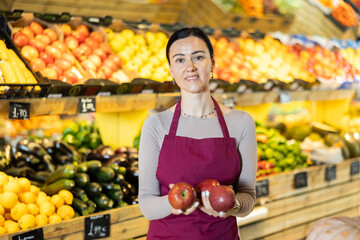 Polite middle-aged female seller offering red apples standing by counter in large grocery store