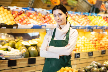 Young woman seller in apron offers fresh vegetables and fruits in vegetable shop