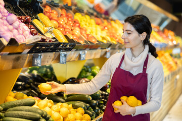 Vegetable shop assistant working in greengrocery. Woman seller in sales area with lemon in hands. Employee demonstrates supply of fruit. Local and imported farm vegetables and fruits