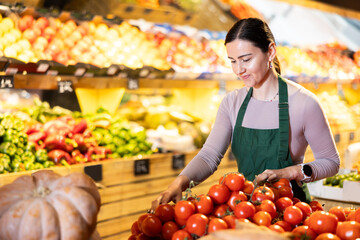 Glad middle-aged female marketer setting tomatoes in market basket in big greengrocery