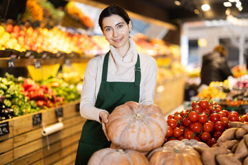 Female vendor stands in the vegetable section of a supermarket and checks a seasonal pumpkin near the counter. Shop employee monitors the quality of products