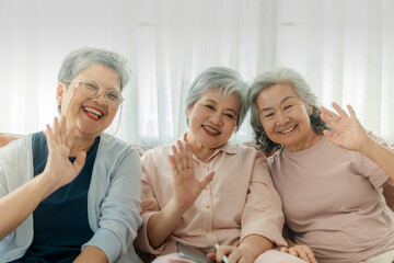 Three elderly women sit together, smiling and waving at the camera, radiating joy and friendship. The image emphasizes companionship, happiness, and togetherness in a relaxed, homey setting.