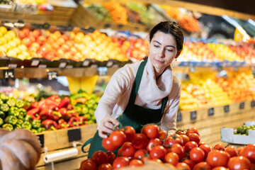 Female seller stands near the counter with red and tomatoes, checks the quality and puts them on the shelf. Supermarket worker keeps an eye on the windows