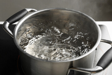 Cooking pot with boiling water and stove on white table against grey background, closeup