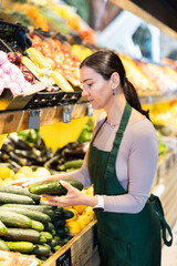 Attentive middle-aged female seller putting cucumbers on stand in fruit and vegetable market