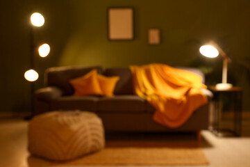 Interior of night living room with sofa, pouf and glowing lamps near green wall, blurred view