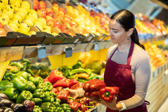 Positive middle-aged female seller putting red peppers on stand in fruit and vegetable market