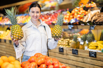 Positive middle-aged woman choosing pineapple standing at counter in large vegetable market