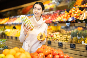 Woman chooses a ripe papaya in a supermarket against the background of shelves with fruit. Customer in the vegetable department looks at exotic fruits