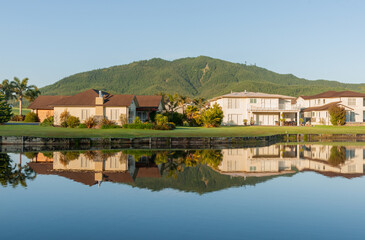 Idyllic residential home reflections in calm waterway foreground and green hill backdrop under blue...