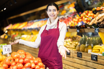 Saleswoman in a red apron stands near the counter with wagons and invites customers to buy quality products in the supermarket