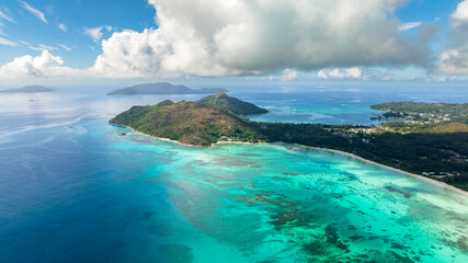 A lush green island stretches along the coast, with colorful coral reefs visible beneath turquoise waters. Praslin, Seychelles.